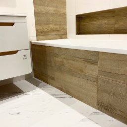 Domowa Przestrzeń Dawid Rogacki - Nowoczesna łazienka z wanną obudowaną panelami imitującymi drewno, białe płytki podłogowe z marmurkowym wzorem i wisząca szafka z drewnianymi uchwytami.