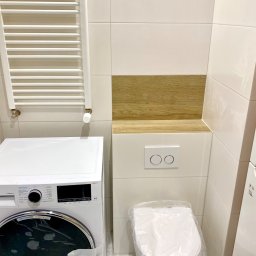 Domowa Przestrzeń Dawid Rogacki - Nowa, biała pralka i toaleta w folii ochronnej w łazience z białymi i drewnopodobnymi płytkami oraz grzejnikiem drabinkowym.