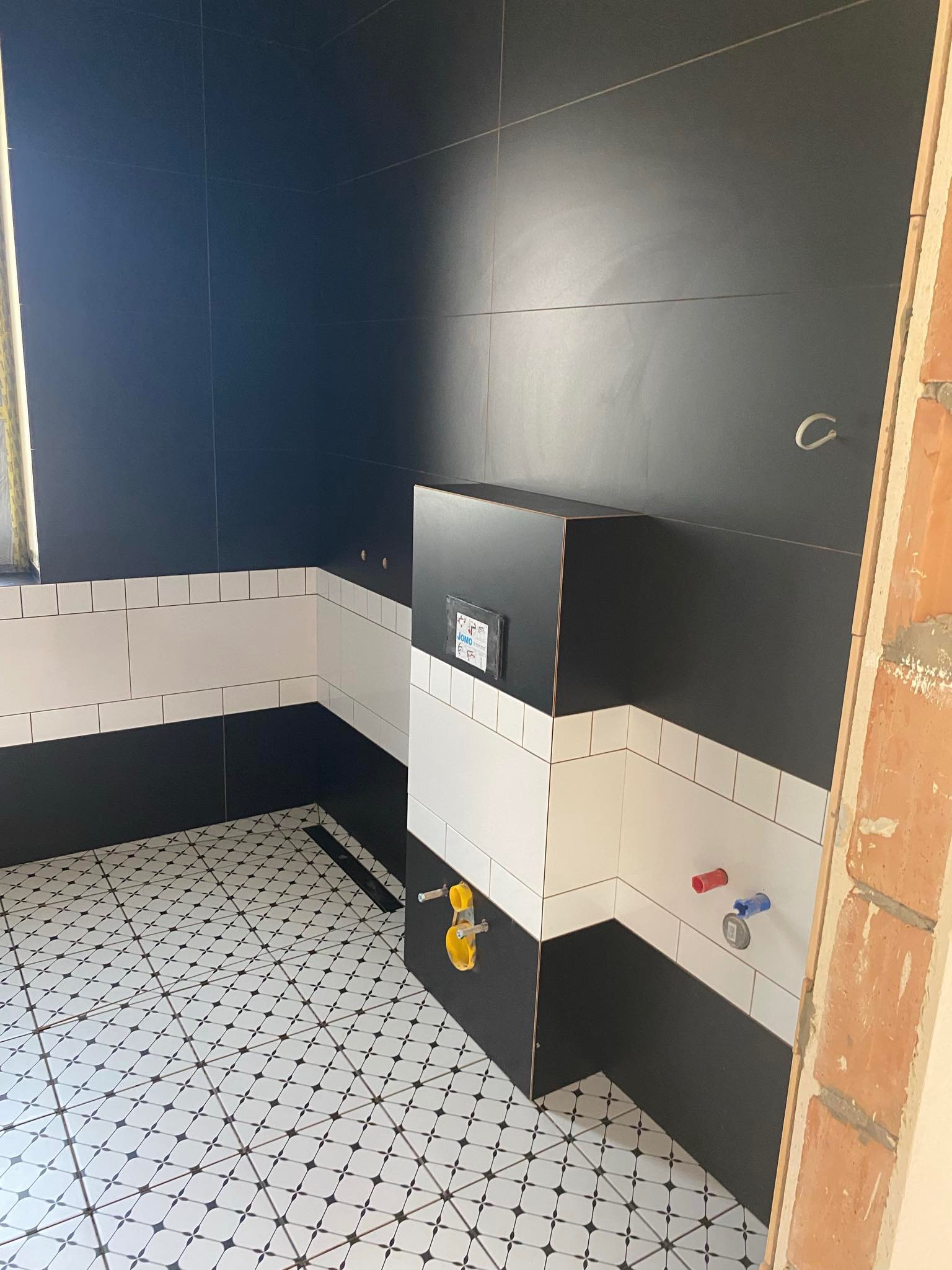 Częściowo wykończona łazienka z czarno-białymi płytkami na ścianach i podłodze w geometryczny wzór, widoczne instalacje hydrauliczne oraz fragment surowej ściany z cegły.