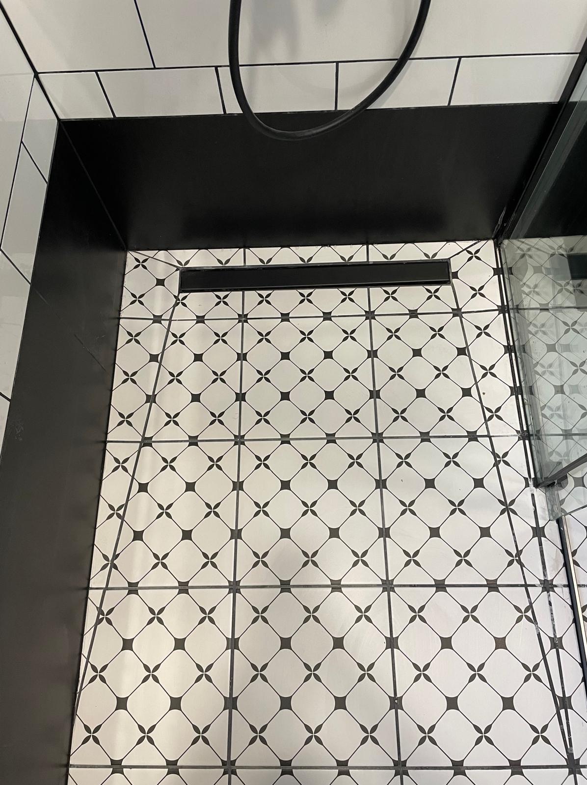 Wzorzyste, biało-czarne płytki podłogowe w kabinie prysznicowej z czarnym odpływem liniowym i czarnym brodzikiem.
