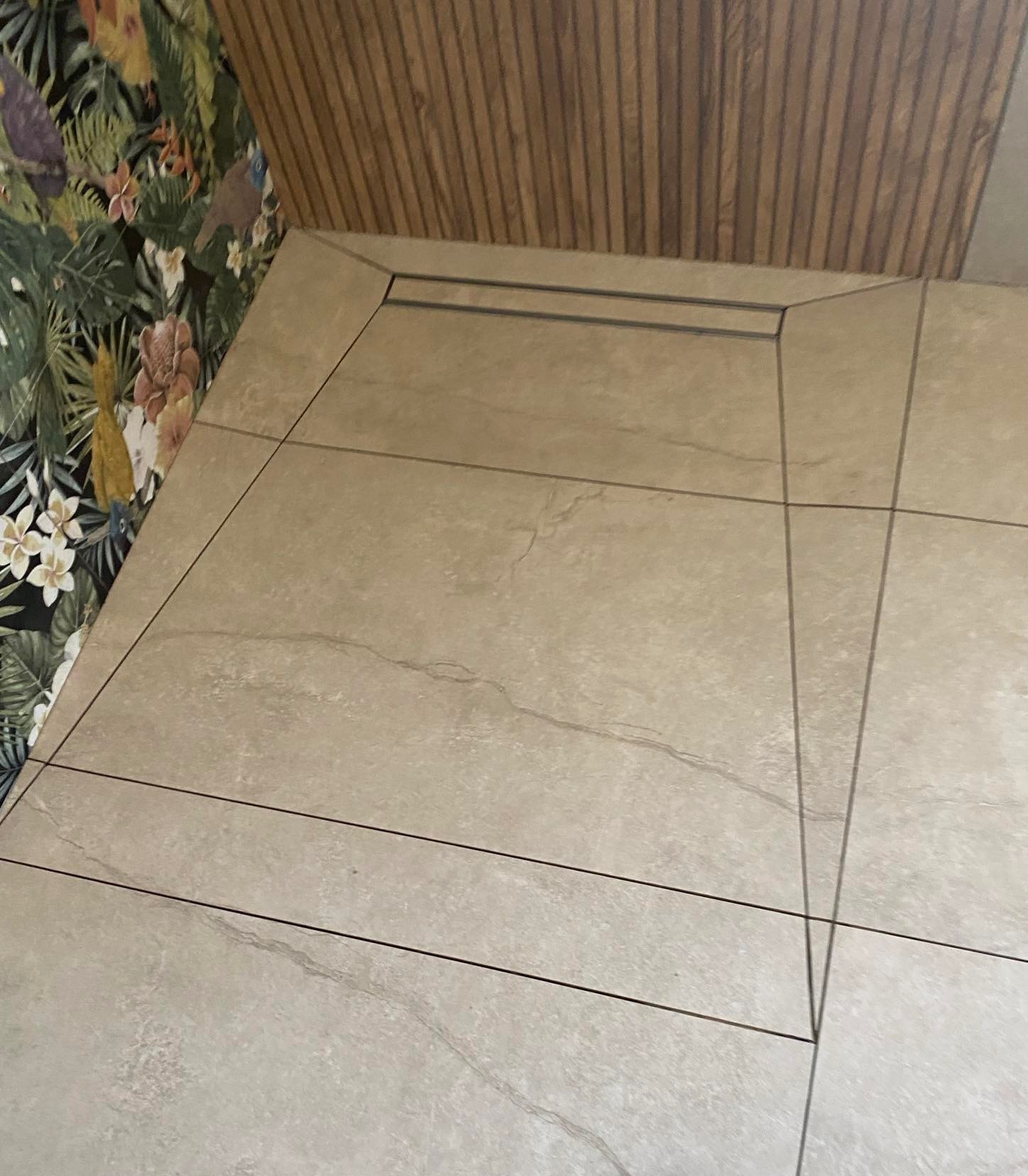 Beżowe płytki podłogowe z delikatnym wzorem żyłkowania, ułożone w regularny wzór, kontrastują z tropikalną tapetą w kwiaty i ptaki oraz drewnianą okładziną ściany.