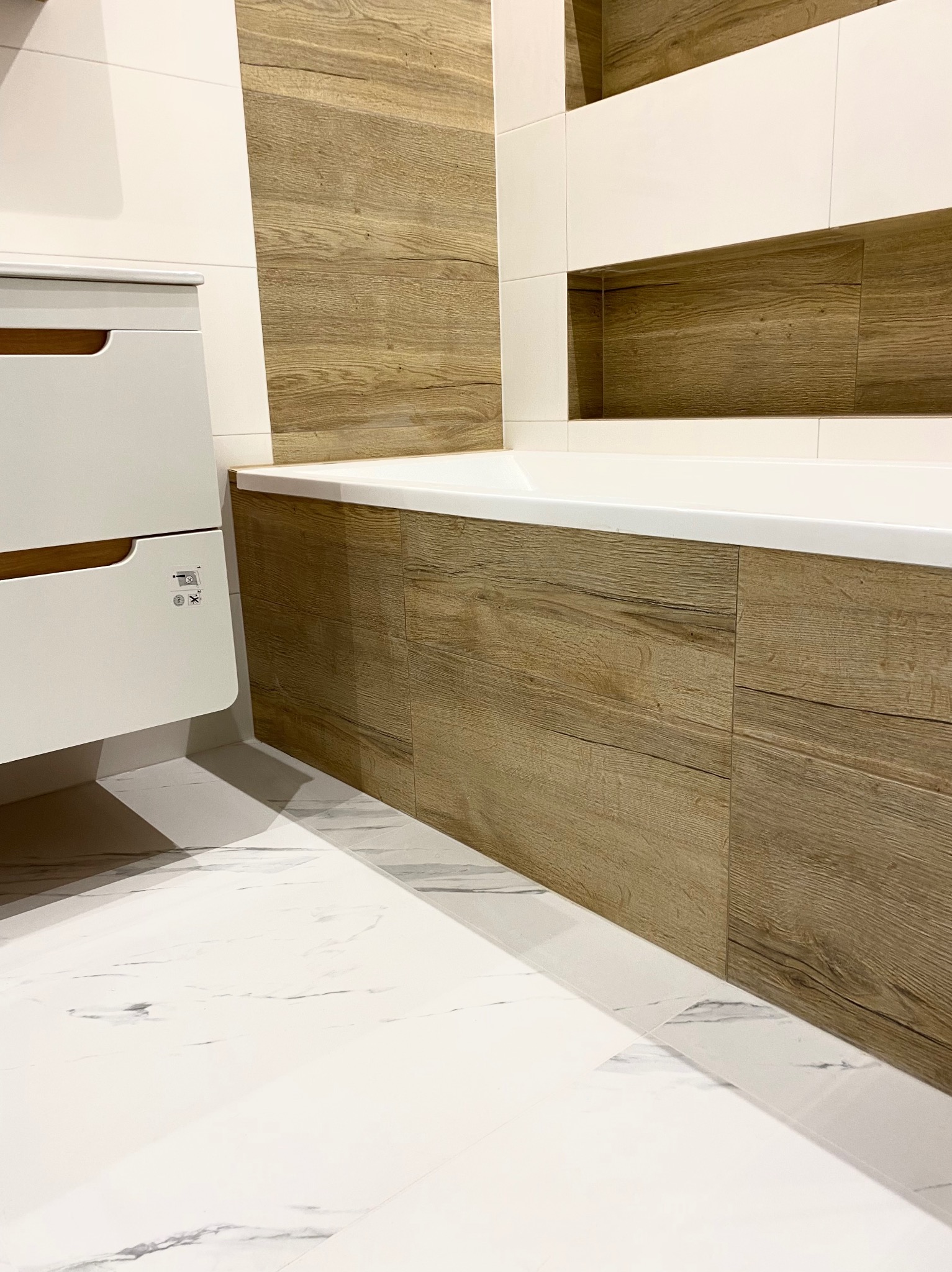 Nowoczesna łazienka z wanną obudowaną panelami imitującymi drewno, białe płytki podłogowe z marmurkowym wzorem i wisząca szafka z drewnianymi uchwytami.