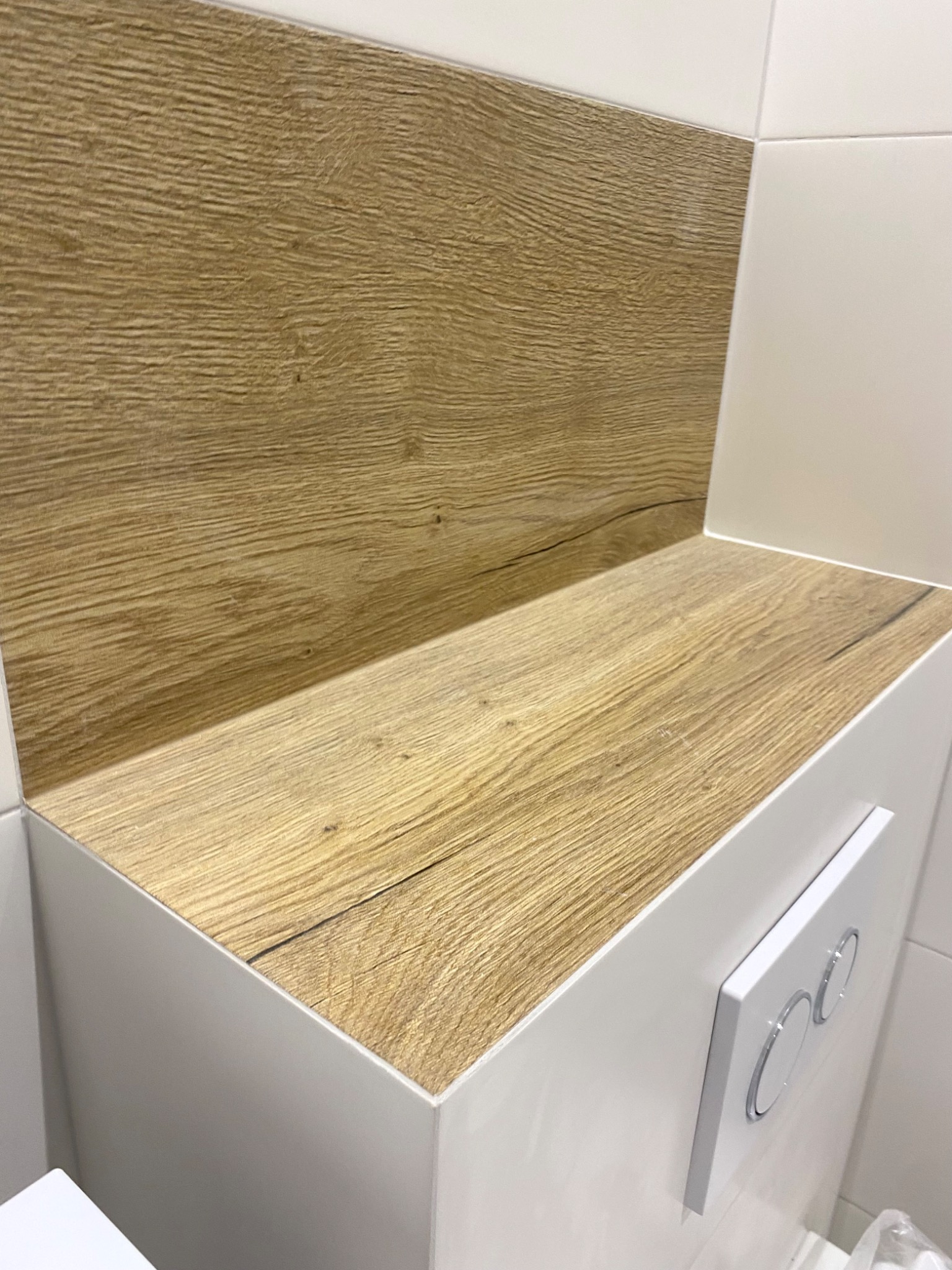 Zabudowa stelaża WC z drewnopodobną półką i białymi płytkami, widoczny przycisk spłukujący.