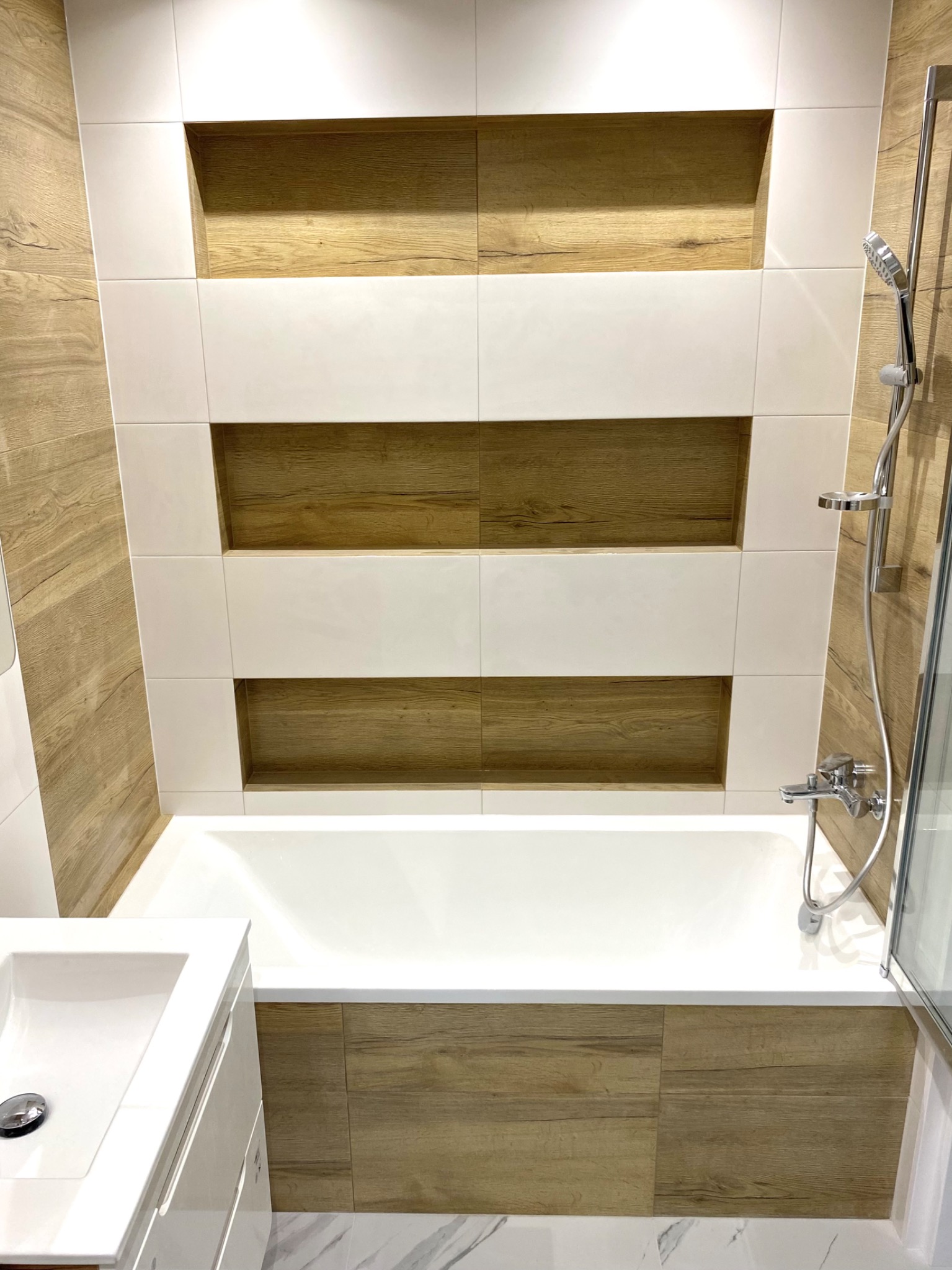 Nowoczesna łazienka z wanną obudowaną płytkami imitującymi drewno, białe płytki z wnękami na ścianie i marmurowa podłoga.