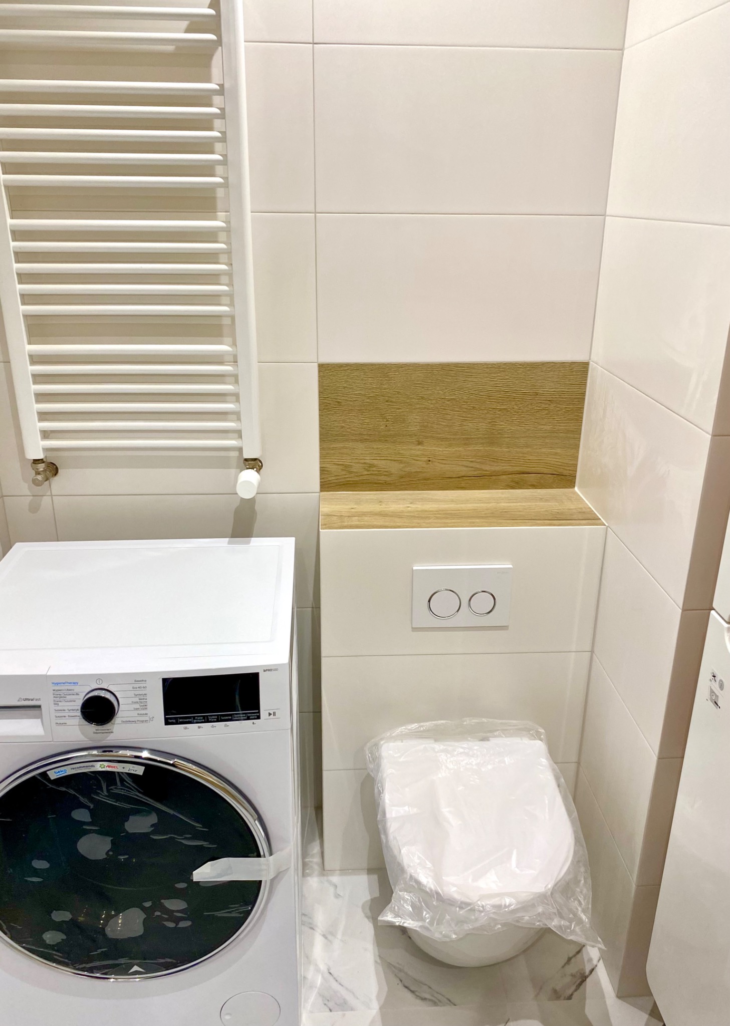 Nowa, biała pralka i toaleta w folii ochronnej w łazience z białymi i drewnopodobnymi płytkami oraz grzejnikiem drabinkowym.