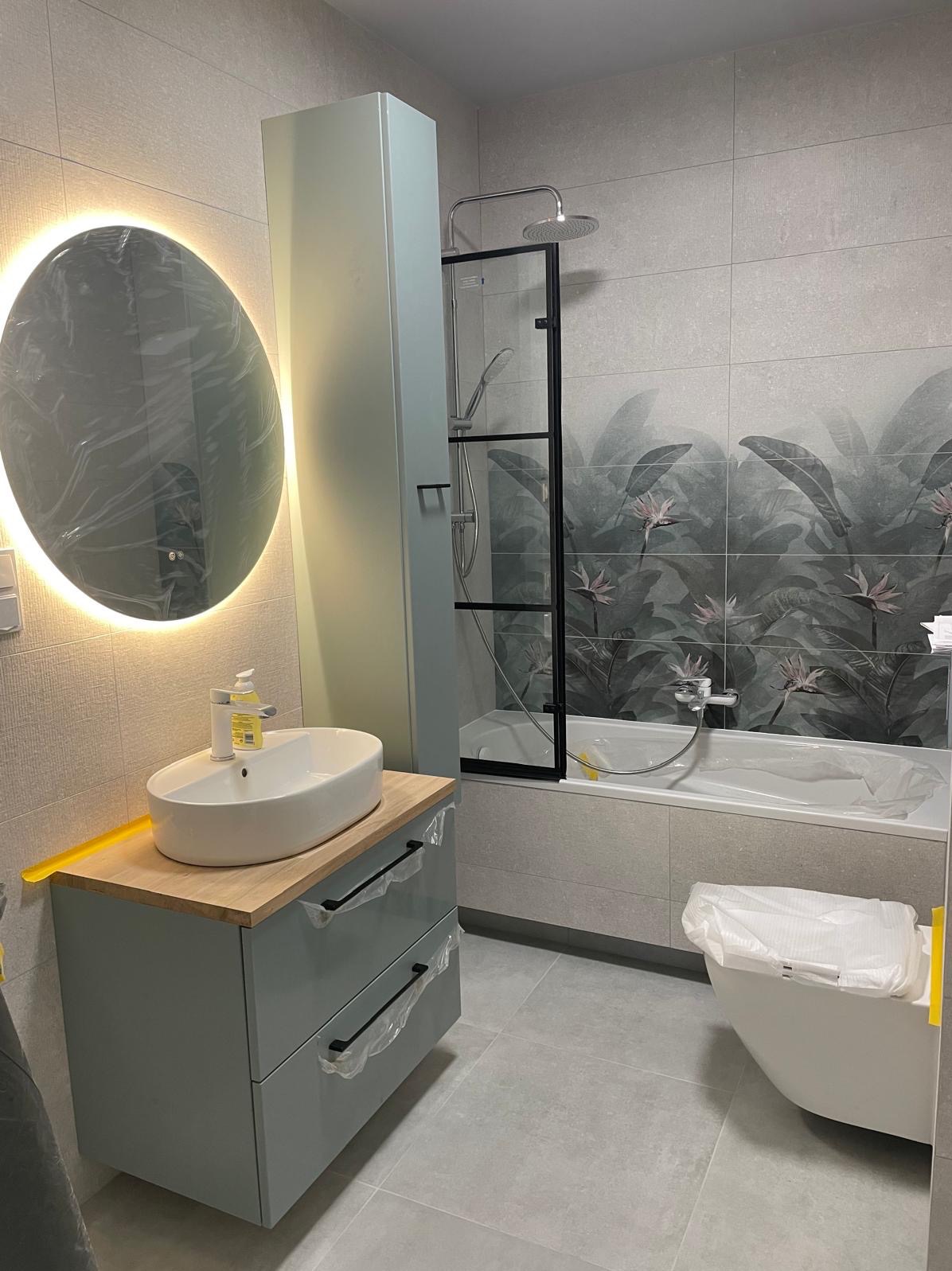 Nowoczesna łazienka po remoncie: szare płytki z motywem roślinnym, umywalka na szafce z drewnianym blatem, okrągłe lustro z podświetleniem LED i kabina prysznicowa.