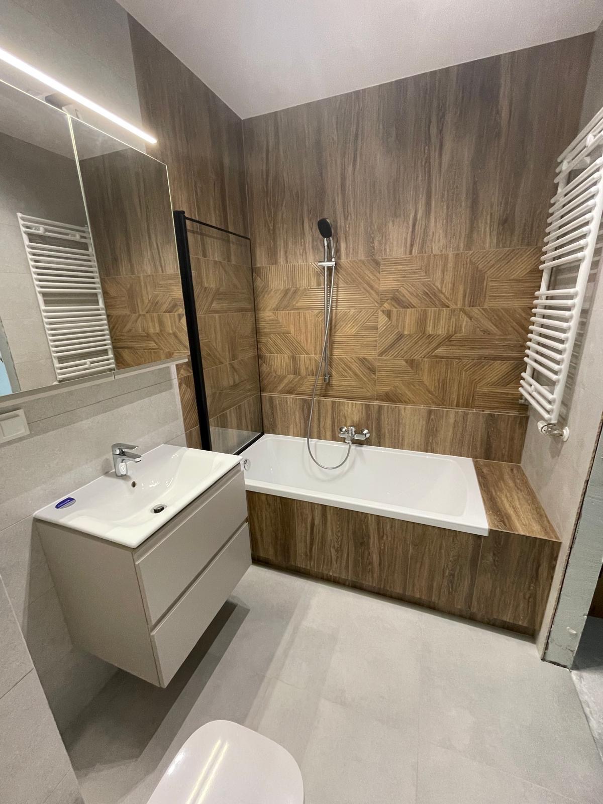 Nowoczesna łazienka po remoncie: drewnopodobne płytki na ścianach, biała wanna z zabudową, umywalka z szafką w odcieniach szarości, grzejnik drabinkowy.