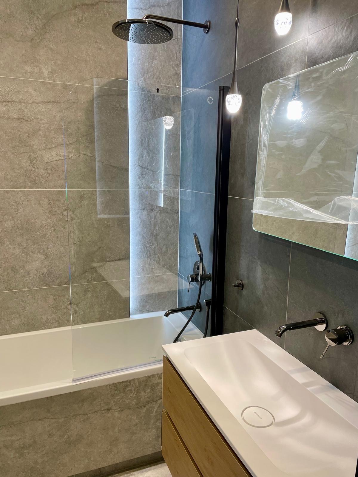 Nowoczesna łazienka z szarą płytką, kabiną prysznicową typu walk-in, białą wanną i umywalką z drewnianą szafką. Minimalistyczne oświetlenie.