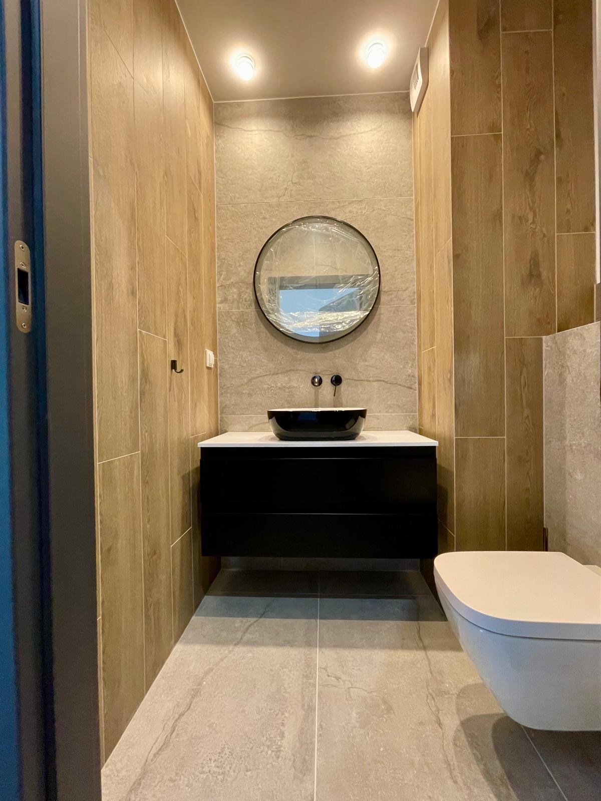 Nowoczesna łazienka z drewnopodobnymi płytkami, czarną szafką z umywalką i okrągłym lustrem w folii ochronnej. Minimalistyczny design, neutralna kolorystyka.
