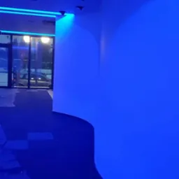 Nowoczesne wnętrze z zakrzywioną białą ścianą podświetloną na niebiesko, widoczne reflektory sufitowe i przeszklone wejście.