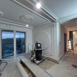 Mix-Bud Świecie usługi remontowo - budowlane - Wnętrze w trakcie prac wykończeniowych: świeżo pomalowane ściany z dekoracyjnymi sztukateriami, zabezpieczona folią część pomieszczenia, widoczny sprzęt malarski i narzędzia na podłodze przy balkonie.