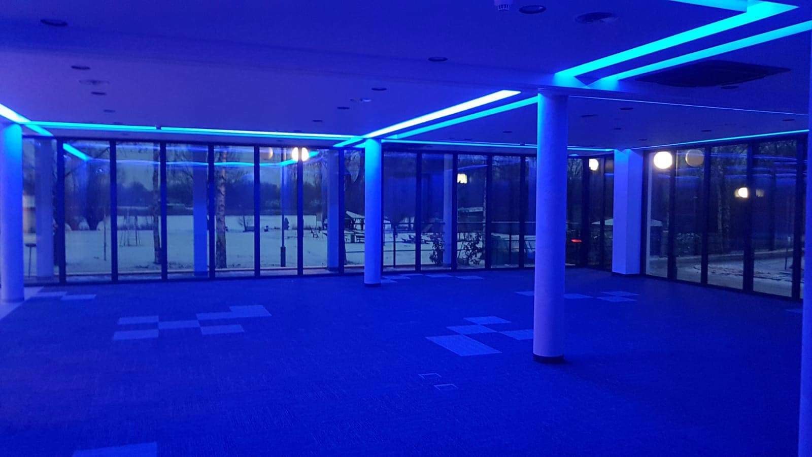 Wnętrze budynku oświetlone intensywnym, niebieskim światłem LED, z widocznymi szklanymi ścianami i kolumnami. Za oknami widoczny zarys drzew i zasnieżony krajobraz.