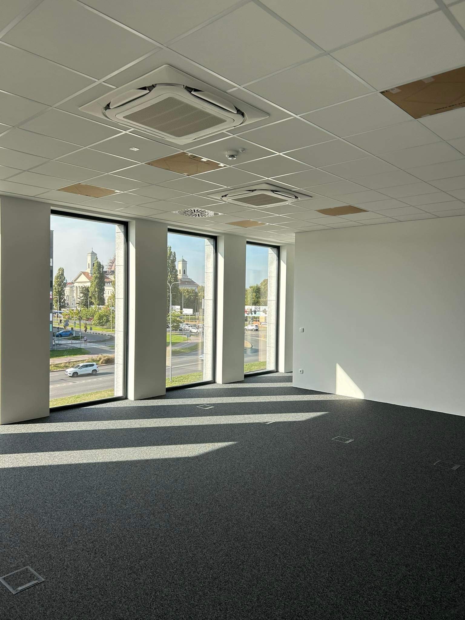 Wnętrze pustego biura z trzema pionowymi oknami, przez które widać ulicę i budynki. Na suficie białe panele i klimatyzatory kasetonowe. Podłoga pokryta szarą wykładziną z kwadratowymi otworami...