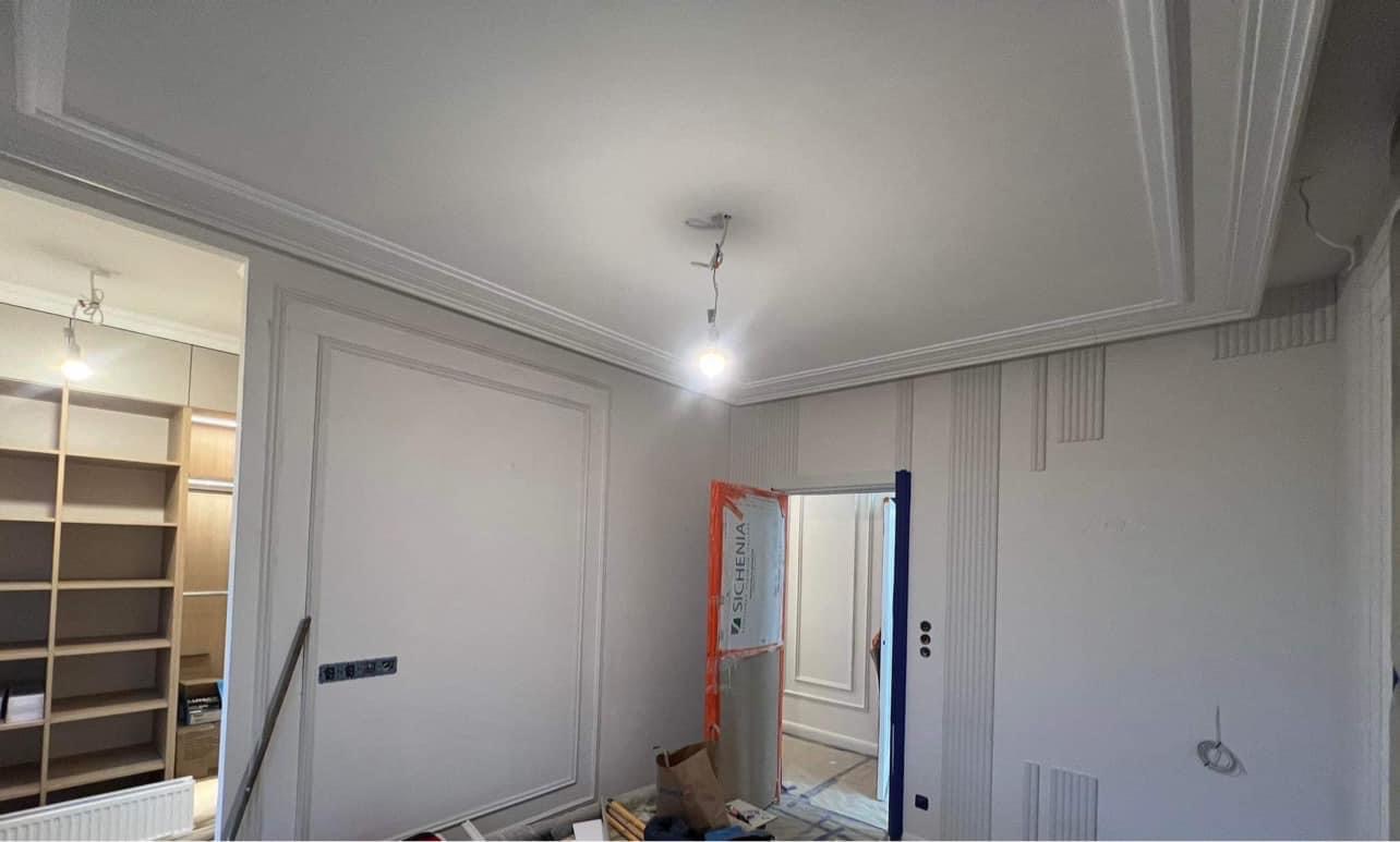 Wnętrze pokoju w trakcie wykańczania, widoczne listwy dekoracyjne na ścianach i suficie, zabezpieczone drzwi folią ochronną, surowa żarówka wisząca na kablu, w tle regał z półkami.