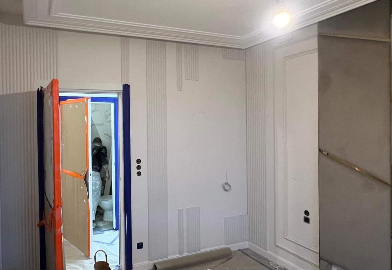 Malowanie dekoracyjne ścian z pionowymi żłobieniami i listwami przysufitowymi w trakcie realizacji, widoczny pracownik w zabezpieczonym pomieszczeniu obok, niebieska taśma malarska zabezpiecza...