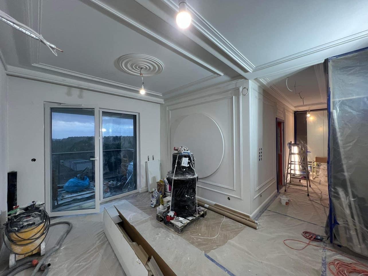 Wnętrze w trakcie prac wykończeniowych: świeżo pomalowane ściany z dekoracyjnymi sztukateriami, zabezpieczona folią część pomieszczenia, widoczny sprzęt malarski i narzędzia na podłodze przy balkonie.