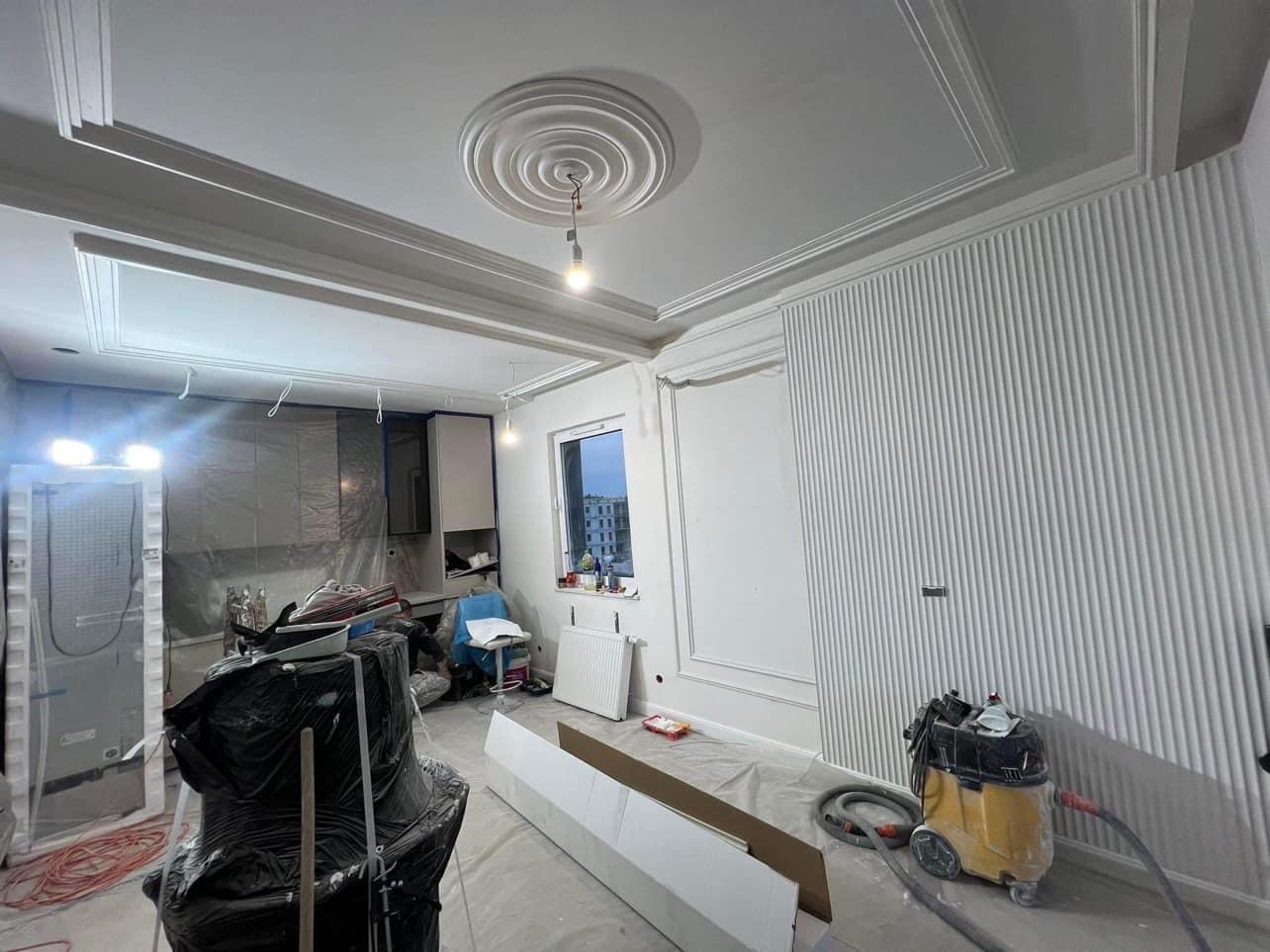 Wnętrze w trakcie wykańczania: białe ściany z dekoracyjnymi panelami pionowymi i listwami przysufitowymi, widoczny odkurzacz przemysłowy i zabezpieczona folią przestrzeń kuchenna.