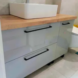 Łazienkowa szafka pod umywalkę z szarymi, lakierowanymi frontami, drewnianym blatem i czarnymi, podłużnymi uchwytami, widoczna biała umywalka nablatowa.