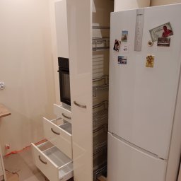 Meble Skrobisz - Biała zabudowa kuchenna w trakcie montażu: wysoka szafka cargo z metalowymi półkami, szafka z piekarnikiem i trzema szufladami z uchwytami, obok biała lodówka z magnesami, podłoga zabezpieczona...