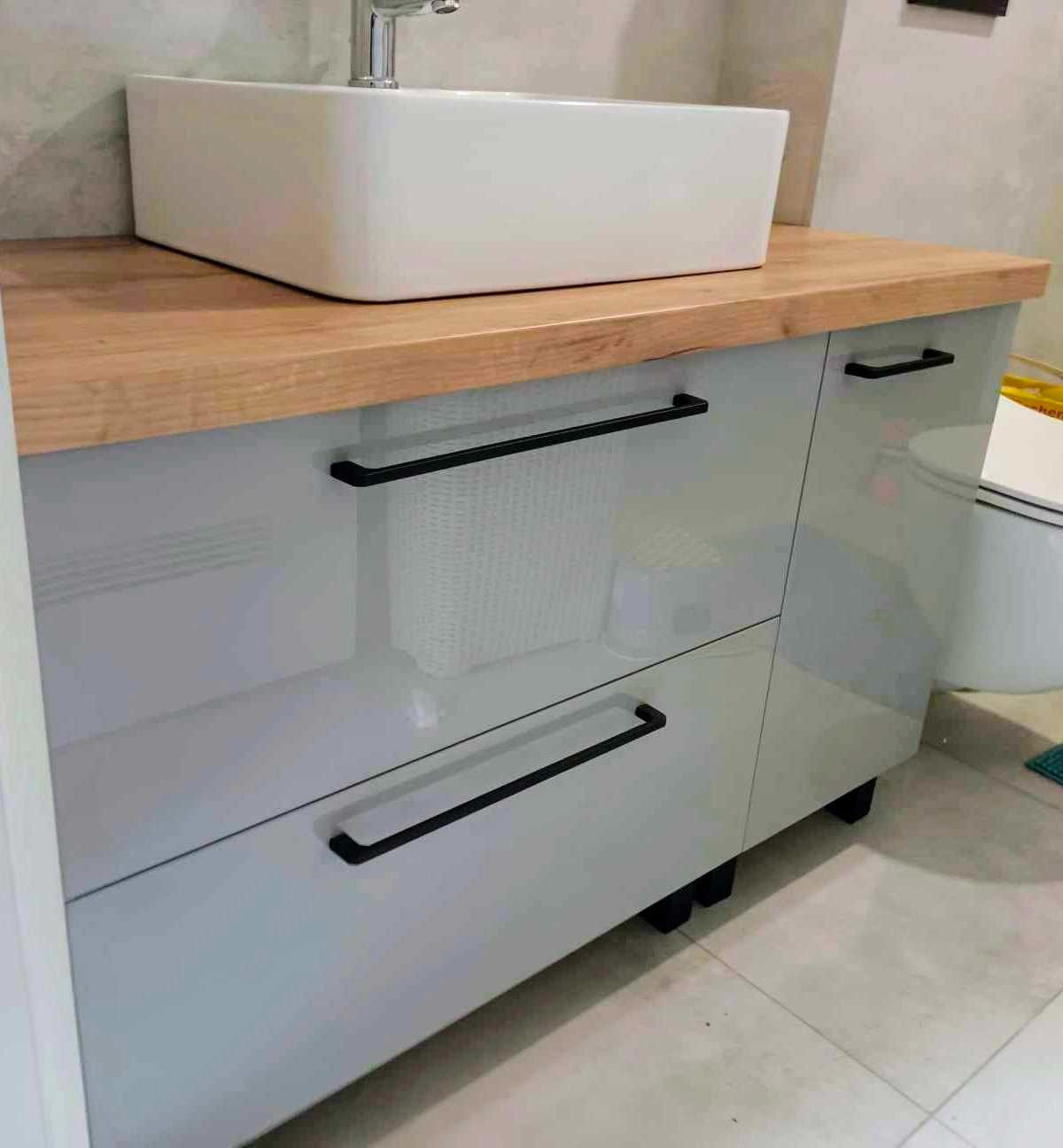 Łazienkowa szafka pod umywalkę z szarymi, lakierowanymi frontami, drewnianym blatem i czarnymi, podłużnymi uchwytami, widoczna biała umywalka nablatowa.