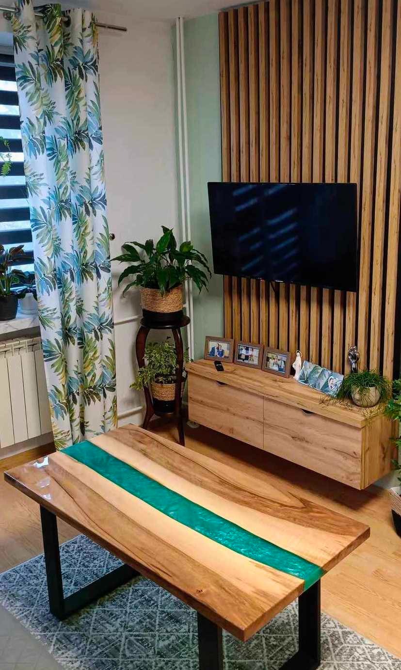 Stół z drewna z żywicą epoksydową w kolorze turkusowym, czarne metalowe nogi, na tle drewnianej ściany z lameli i szafki RTV, dywan w geometryczny wzór.