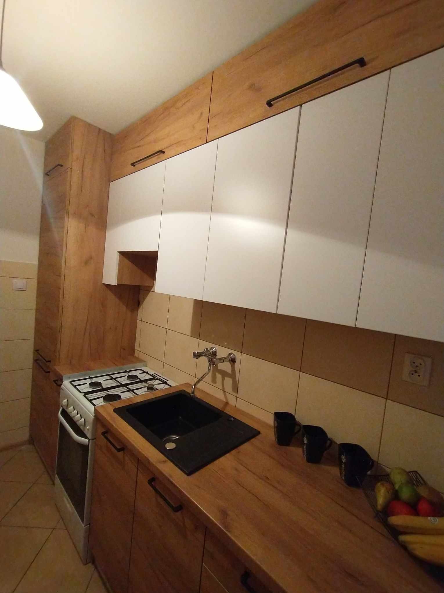 Kuchnia z drewnianymi szafkami dolnymi i białymi górnymi, czarny zlew, biała kuchenka, czarne podłużne uchwyty, owoce w koszyku na blacie