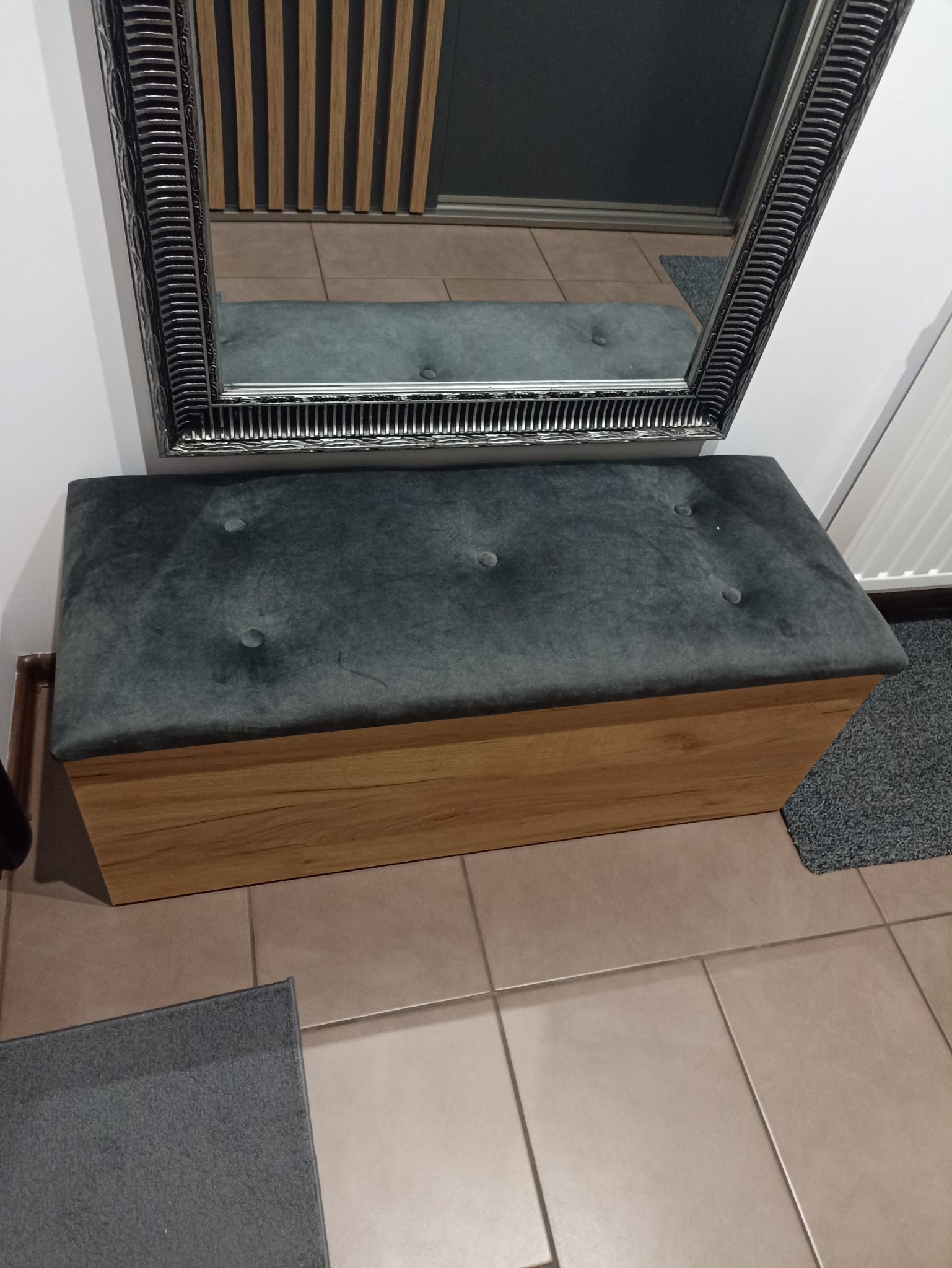 Pufa tapicerowana szarym welurem z drewnianą podstawą, odbicie w lustrze z ozdobną ramą, płytki podłogowe w odcieniach beżu.