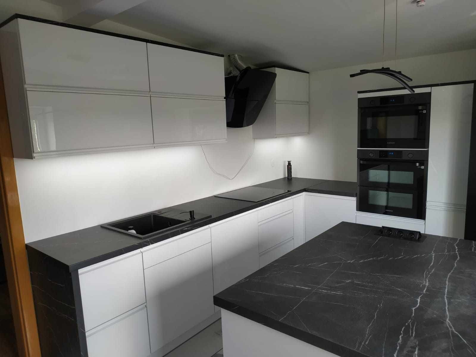 Nowoczesna kuchnia z białymi szafkami i czarnym blatem marmurkowym, czarny zlew, płyta indukcyjna i zabudowane piekarniki Samsung, oświetlenie LED pod szafkami wiszącymi.