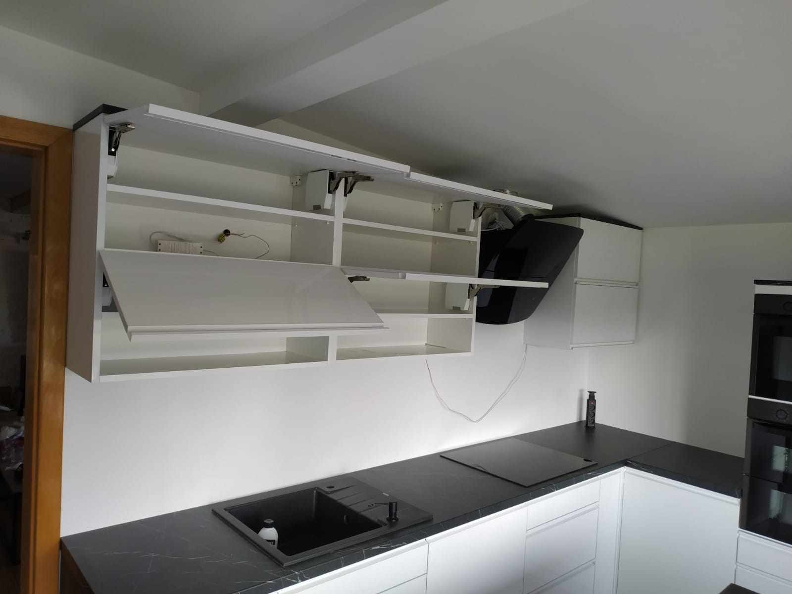 Biała kuchnia z czarnym blatem, szafki górne otwierane do góry z widocznymi mechanizmami, czarny okap i zlew, oświetlenie LED pod szafkami.