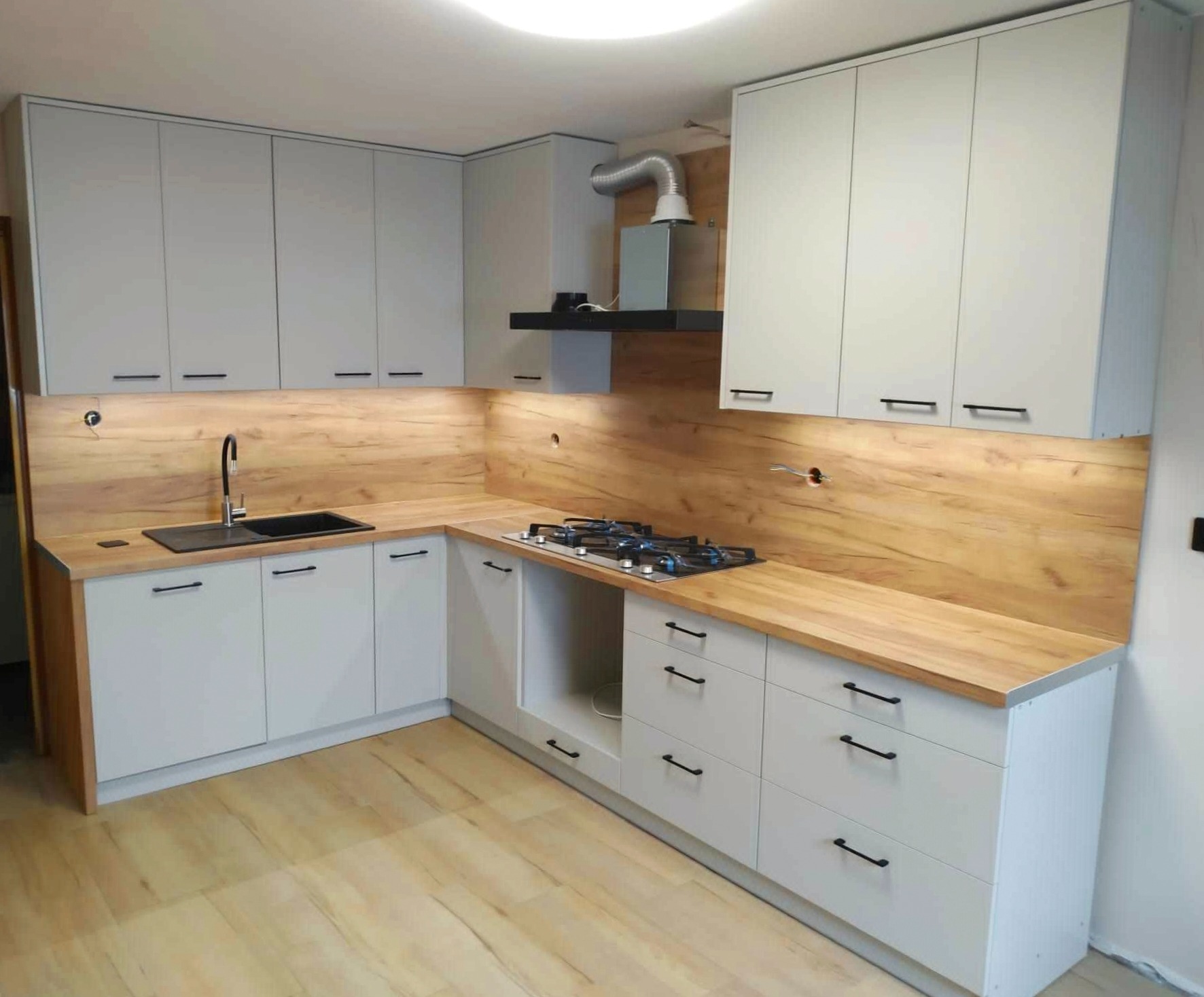 Jasnoszara kuchnia z drewnianym blatem i panelem ściennym, czarny zlewozmywak i bateria, płyta gazowa, oświetlenie podszafkowe