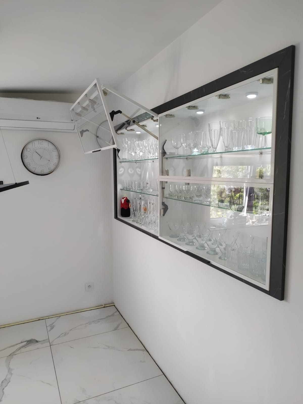 Szklana szafka wnękowa z czarną marmurową ramą, wypełniona różnymi szklankami i kieliszkami, z otwartą górną klapą i oświetleniem LED, zamontowana na białej ścianie z widocznym zegarem i klimatyzacją.