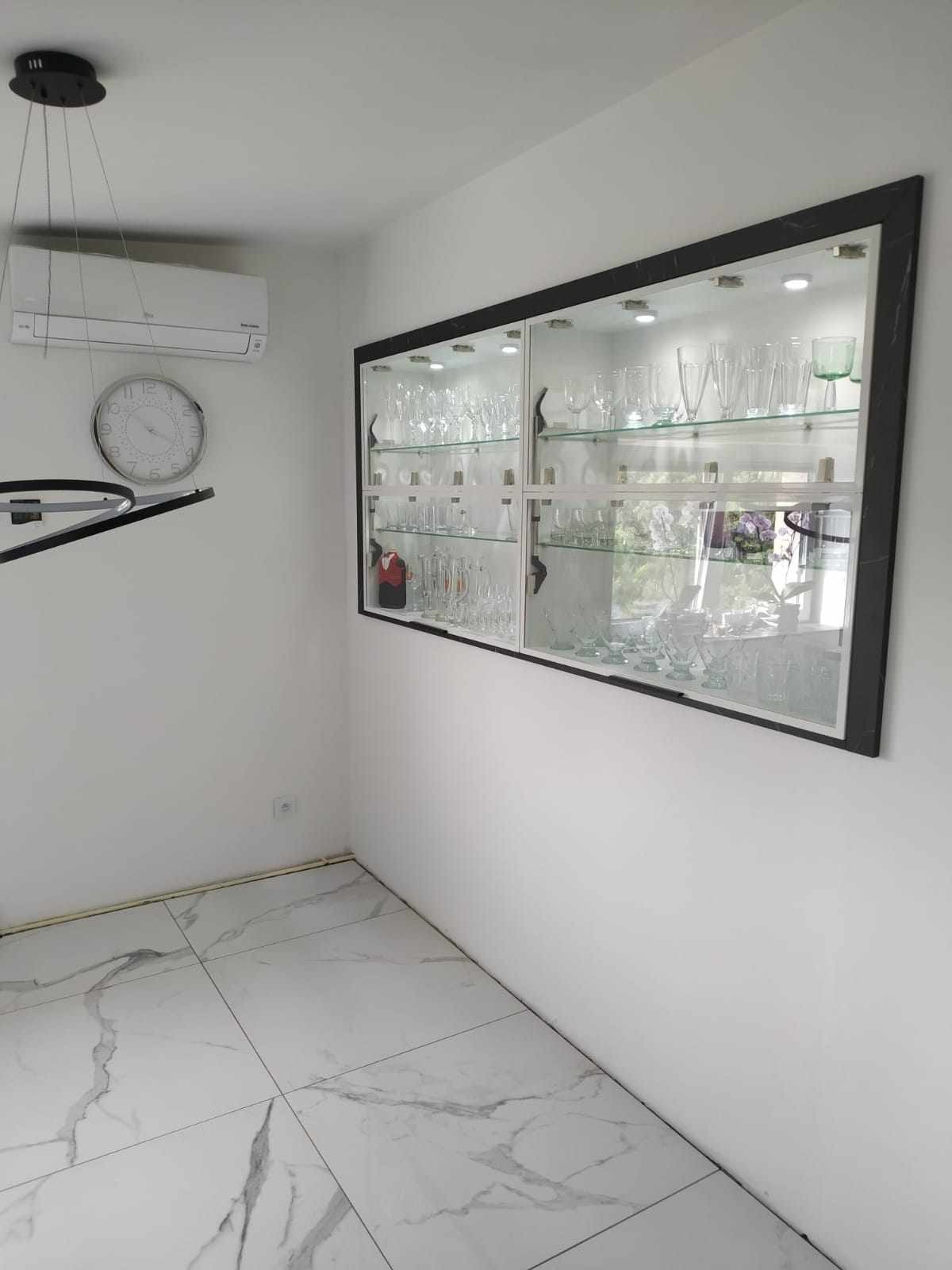 Szklana witryna w czarnej ramie, wbudowana w ścianę, oświetlona od wewnątrz, wypełniona kolekcją szkła, w tle białe ściany, marmurowa podłoga i klimatyzator.