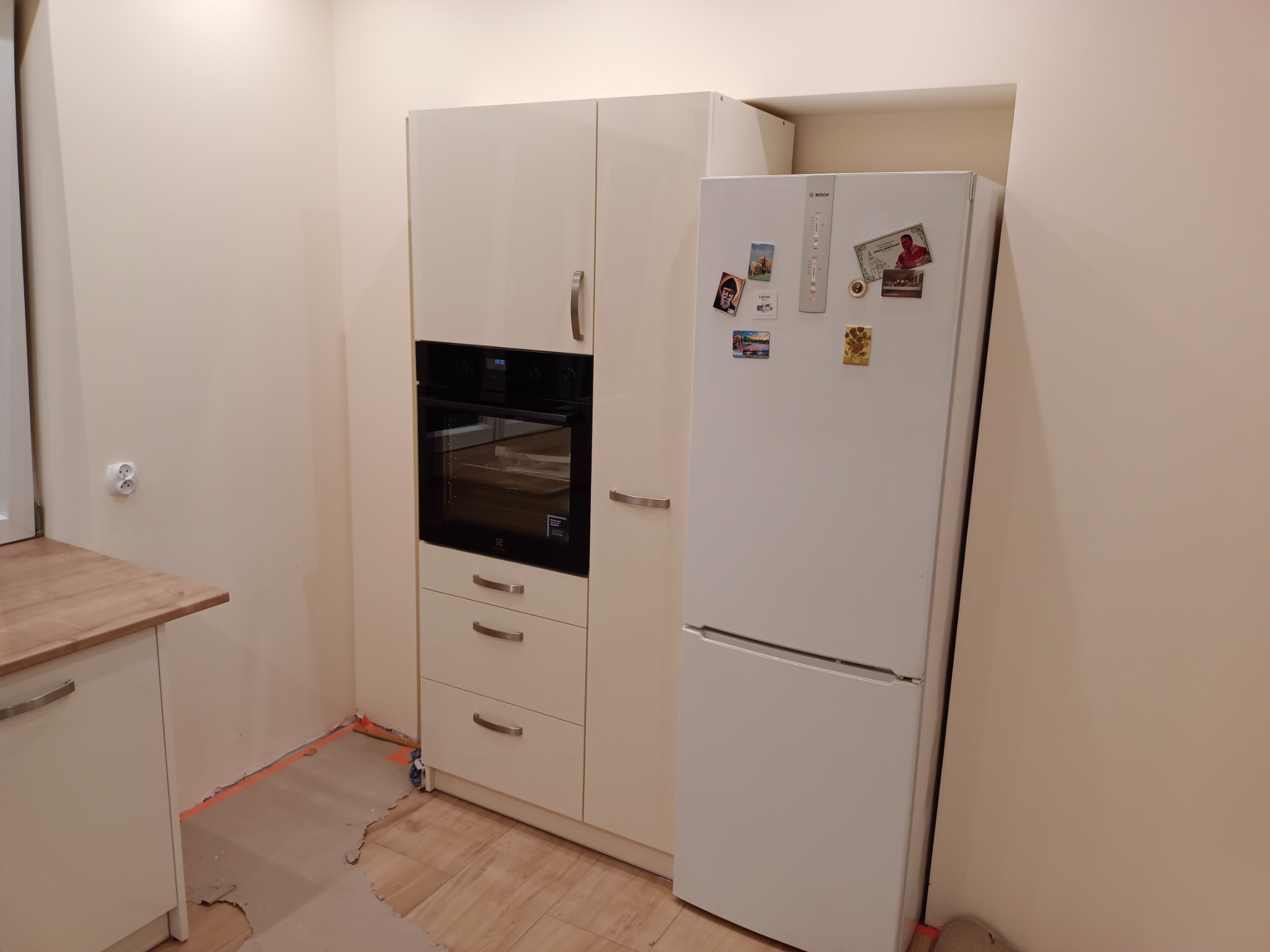 Biała zabudowa kuchenna z piekarnikiem i lodówką w trakcie montażu, widoczne ślady po remoncie na podłodze.