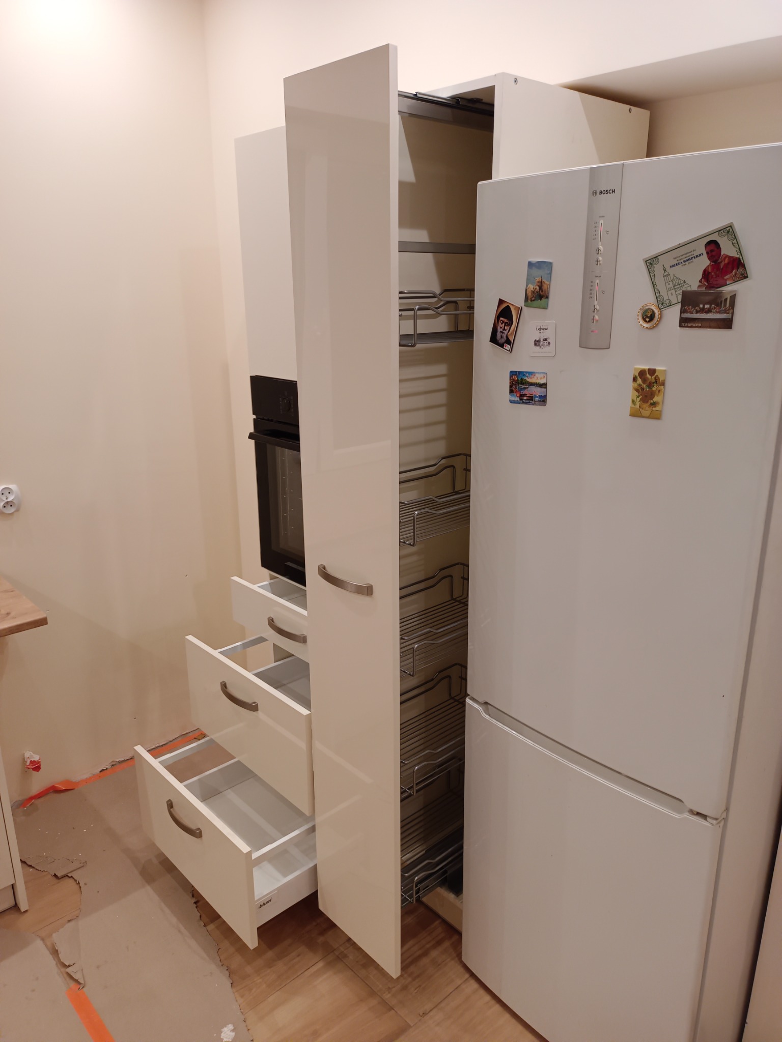 Biała zabudowa kuchenna w trakcie montażu: wysoka szafka cargo z metalowymi półkami, szafka z piekarnikiem i trzema szufladami z uchwytami, obok biała lodówka z magnesami, podłoga zabezpieczona...