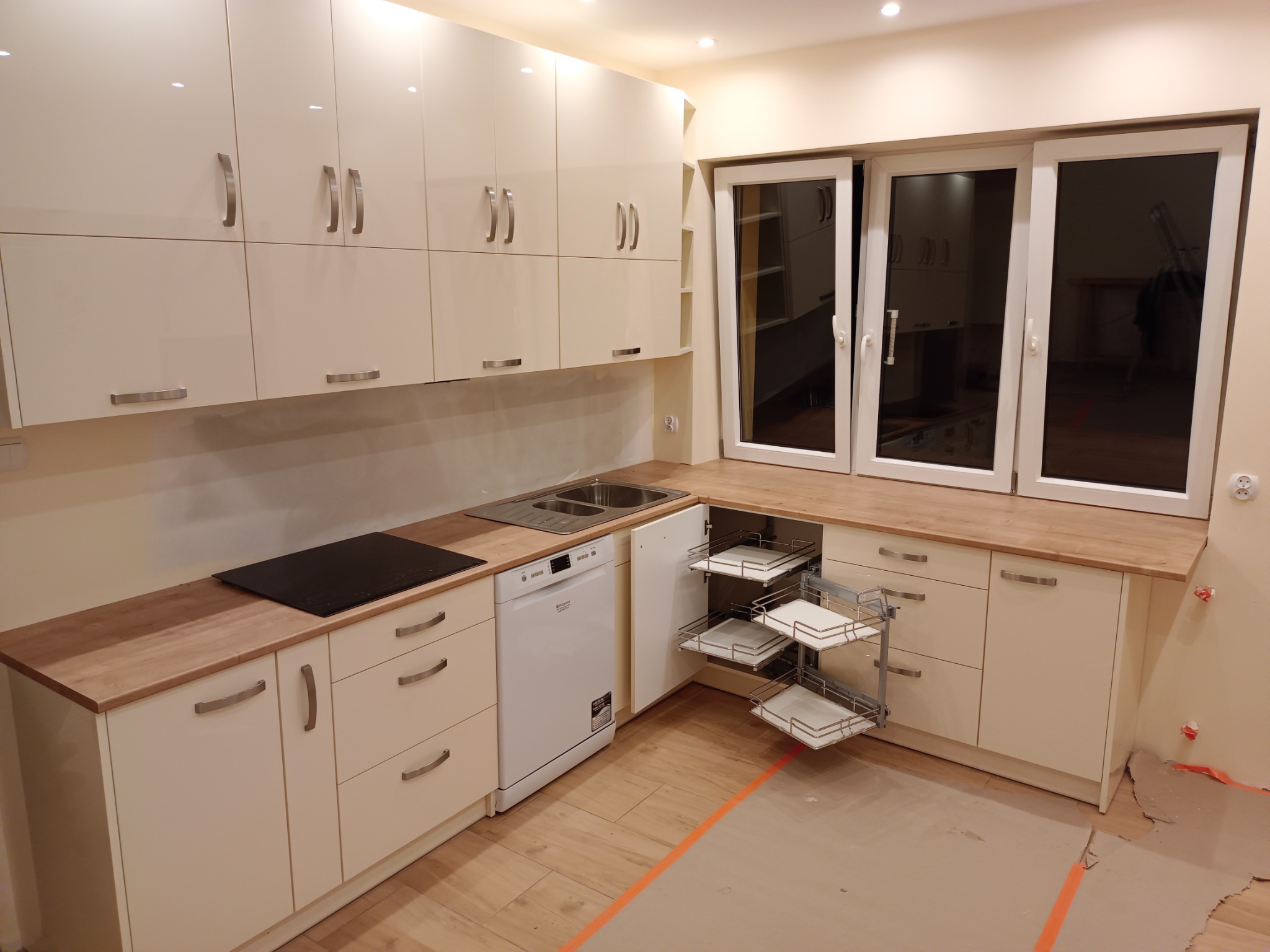 Jasna kuchnia z białymi, lakierowanymi frontami, drewnianym blatem, wysuwanymi półkami cargo i płytą indukcyjną, widoczne niedokończone prace wykończeniowe.