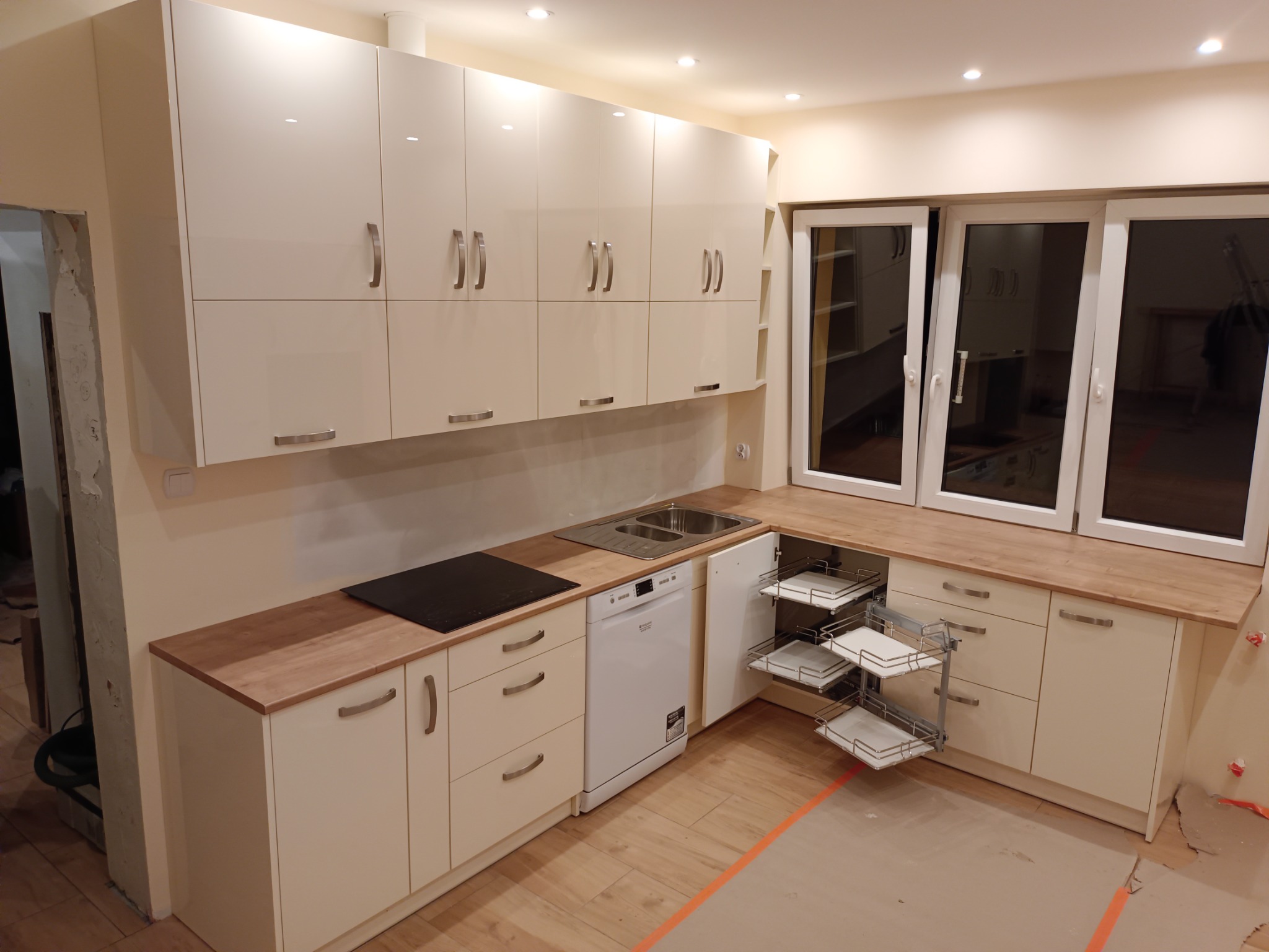 Jasna kuchnia z białymi szafkami na wymiar, drewnianym blatem, wysuwanymi koszami cargo i płytą indukcyjną, w trakcie montażu, z widocznymi śladami prac wykończeniowych na podłodze.