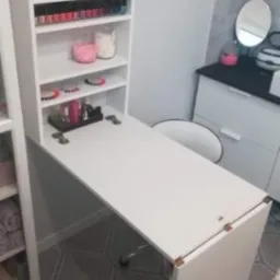 Biała, składana szafka z blatem pełniącym funkcję biurka, z półkami wypełnionymi lakierami do paznokci i akcesoriami kosmetycznymi, ustawiona w pomieszczeniu z jasnymi ścianami i podłogą...