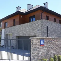 Dwukondygnacyjny dom z elewacją z jasnej cegły i drewnianą górną częścią, widoczny zza metalowej bramy w słoneczny dzień.