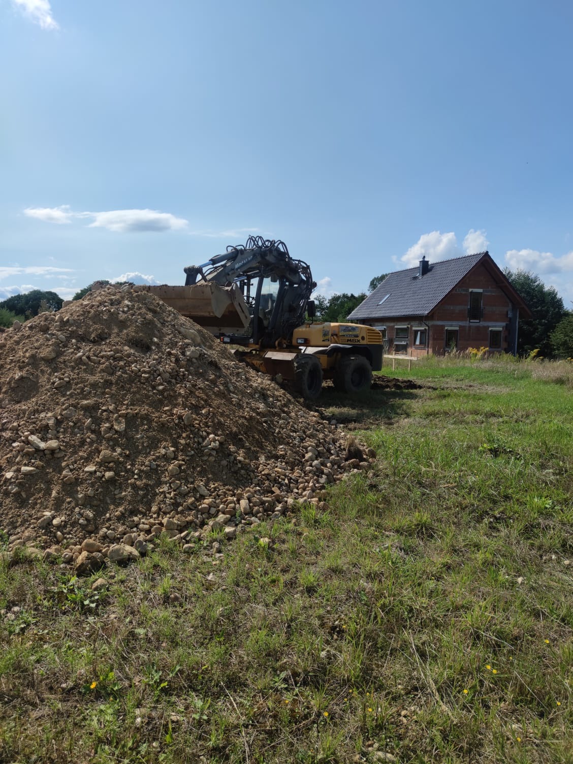 Żółta koparka kołowa stoi obok pryzmy ziemi z kamieniami na tle budującego się domu z czerwonej cegły i błękitnego nieba.