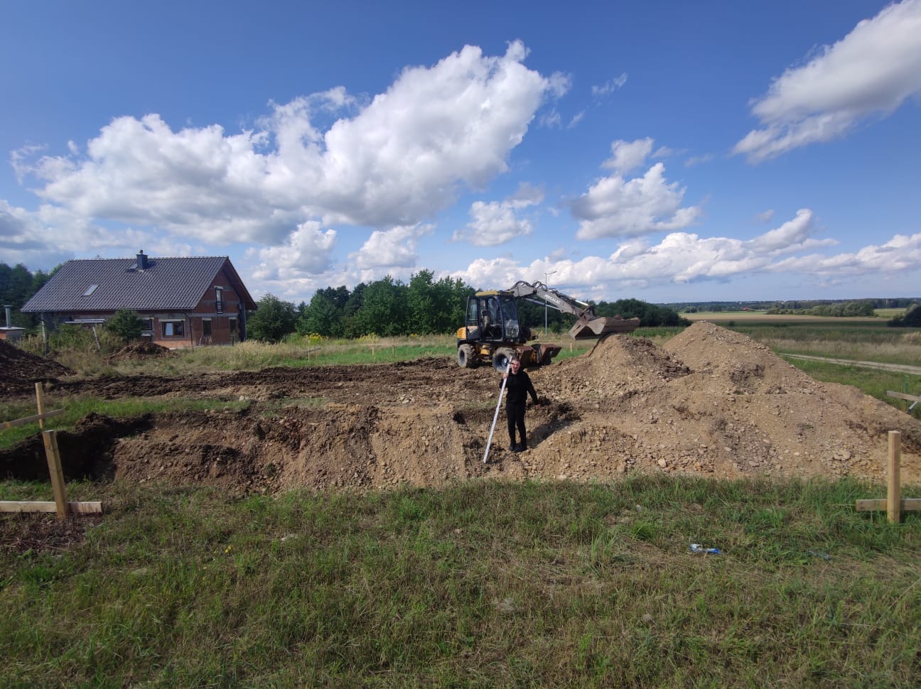 Pracownik z łatą niwelacyjną na tle wykopu pod budowę domu, w tle koparko-ładowarka i dom jednorodzinny pod błękitnym niebem z białymi chmurami.