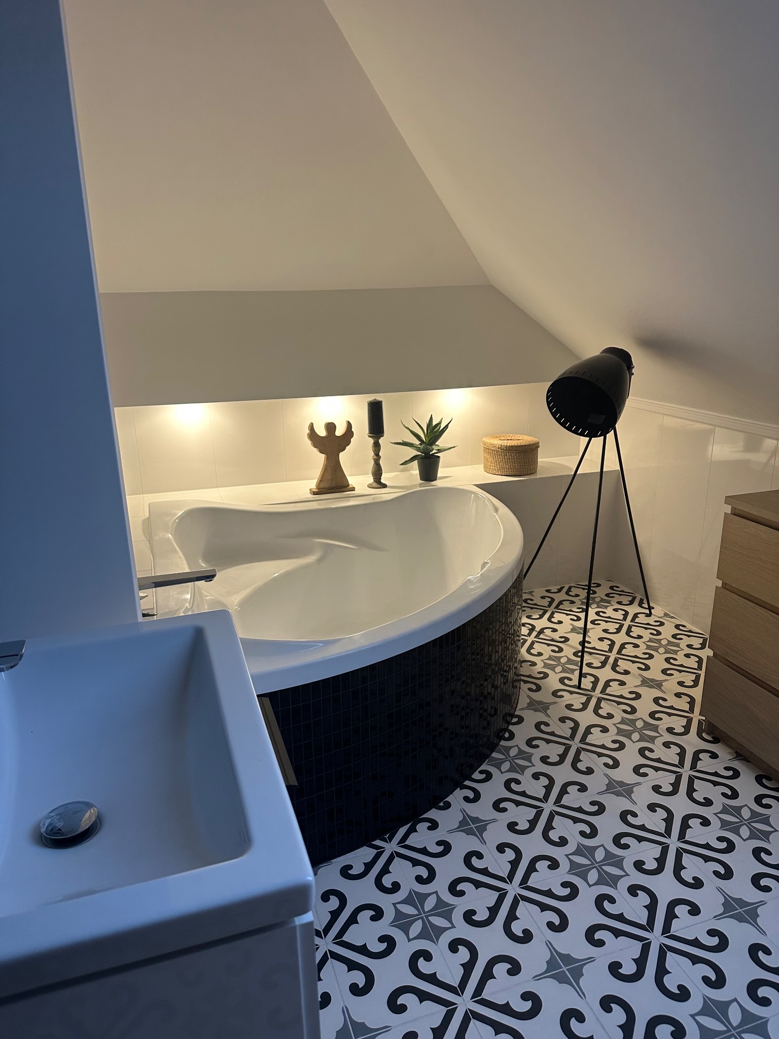 Nowoczesna łazienka na poddaszu z wolnostojącą wanną obudowaną czarną mozaiką, wzorzystą podłogą, oświetleniem LED w niszy i czarną lampą na trójnogu.