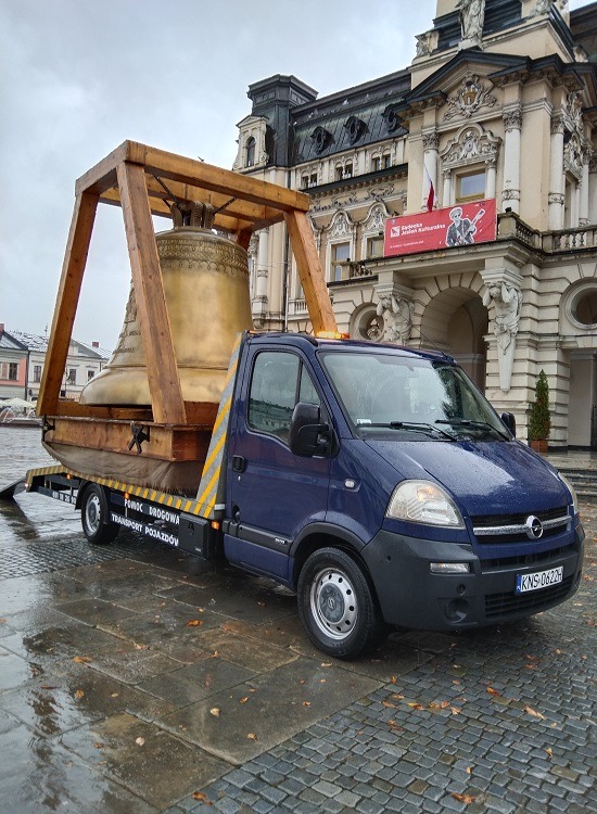 Niebieska laweta z drewnianą konstrukcją przewożąca duży, brązowy dzwon na tle zabytkowego budynku z plakatem.