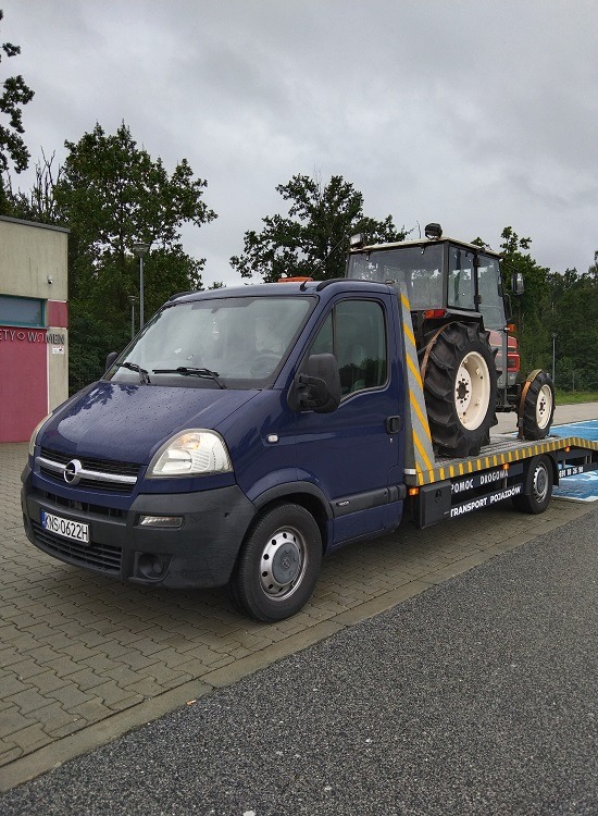 Niebieska laweta z napisem 'Pomoc Drogowa' przewozi traktor z kabiną na parkingu z kostki brukowej w pochmurny dzień.