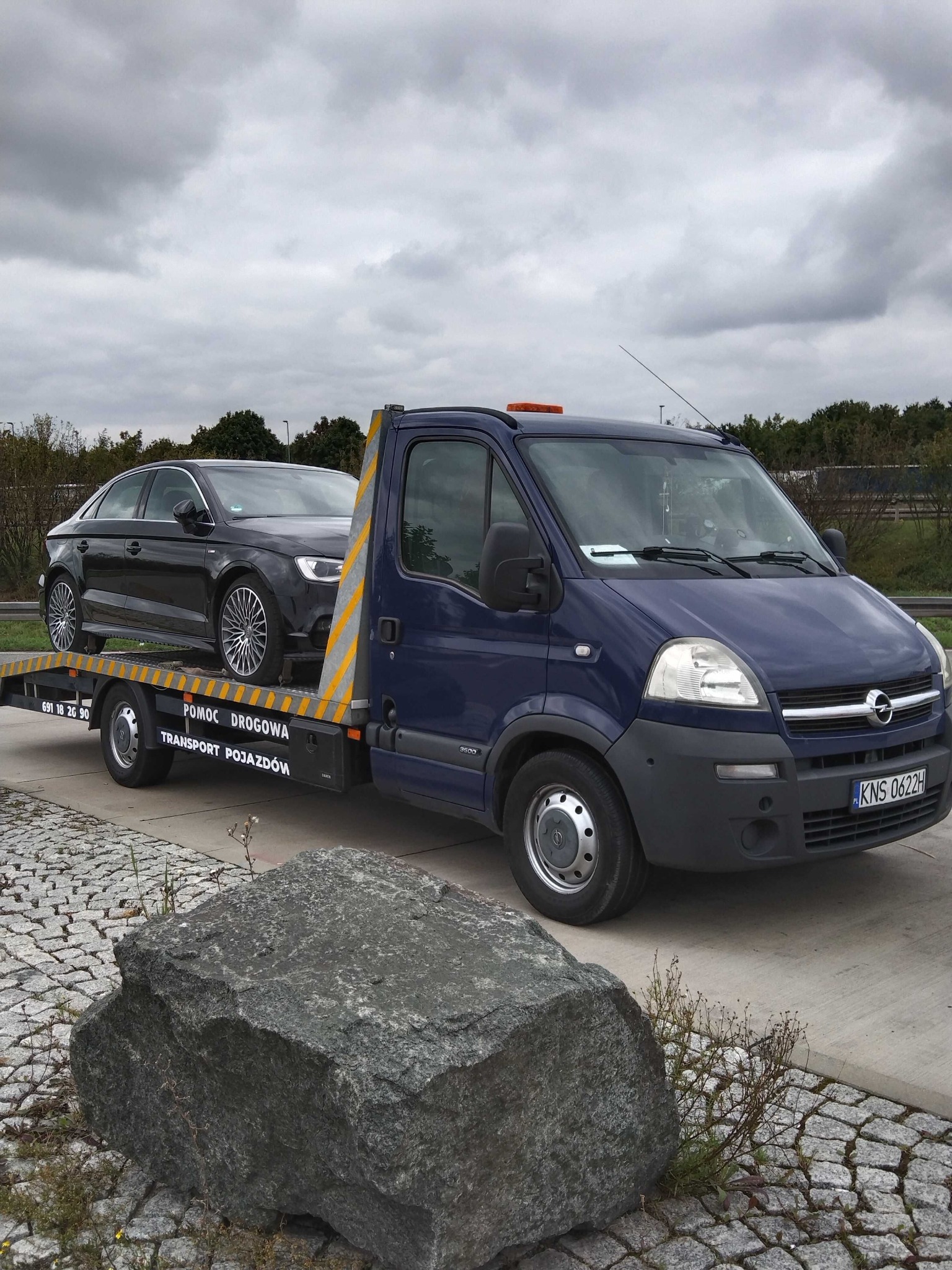 Niebieska laweta Opel z czarnym samochodem osobowym marki Audi na platformie, zaparkowana na kostce brukowej, widoczny duży kamień w dolnym rogu kadru.