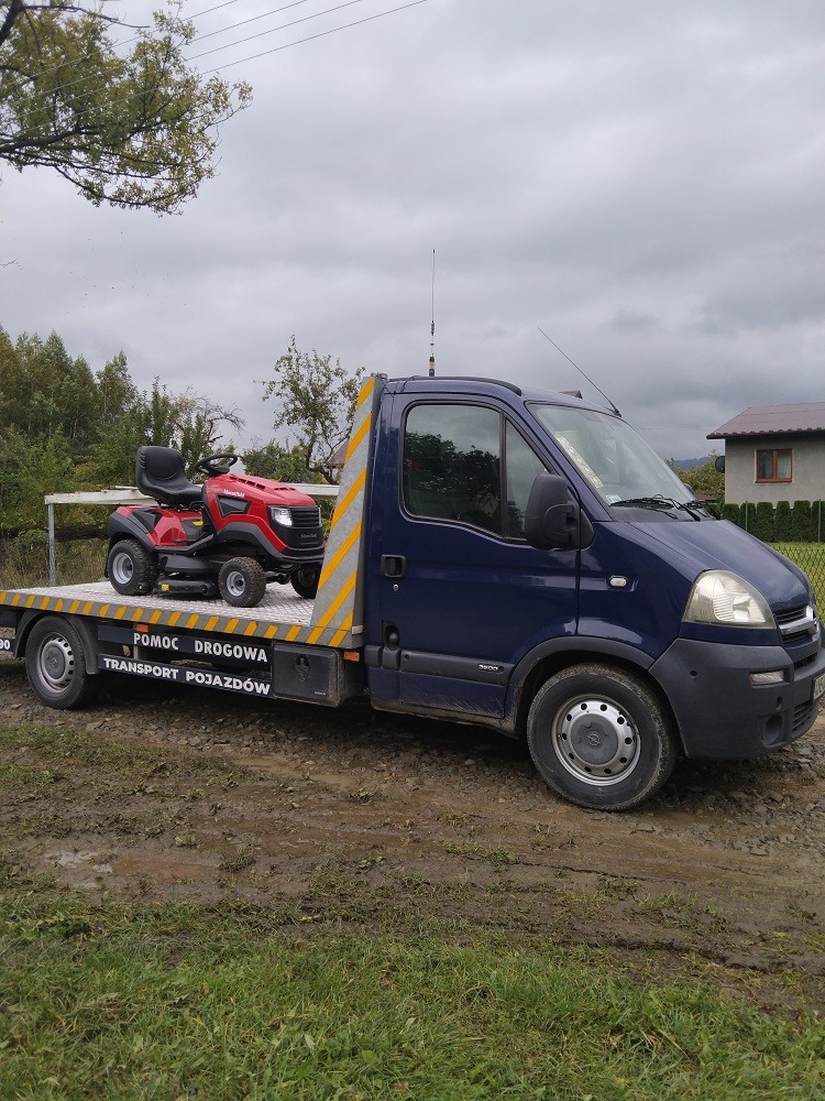 Niebieska laweta z czerwoną kosiarką traktorkiem na platformie, zaparkowana na błotnistym terenie, widoczne napisy 'POMOC DROGOWA TRANSPORT POJAZDÓW'.