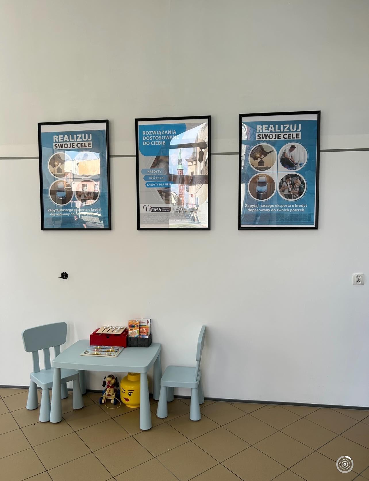 Poczekalnia z dziecięcym stolikiem i krzesłami, na ścianie wiszą oprawione plakaty z hasłem 'Realizuj swoje cele' i logo firmy finansowej oferującej kredyty i pożyczki.