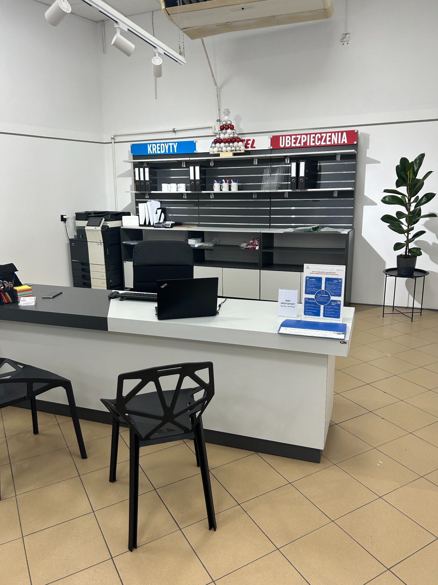 Wnętrze biura z napisem 'Ubezpieczenia' nad regałem, biurko z laptopem i dokumentami, oraz dekoracja świąteczna z bombek.