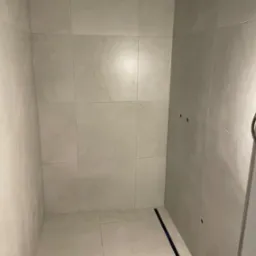 Wykończony narożnik kabiny prysznicowej z jasnymi, marmurkowymi płytkami ceramicznymi na ścianach i podłodze, widoczny odpływ liniowy oraz otwory na armaturę.