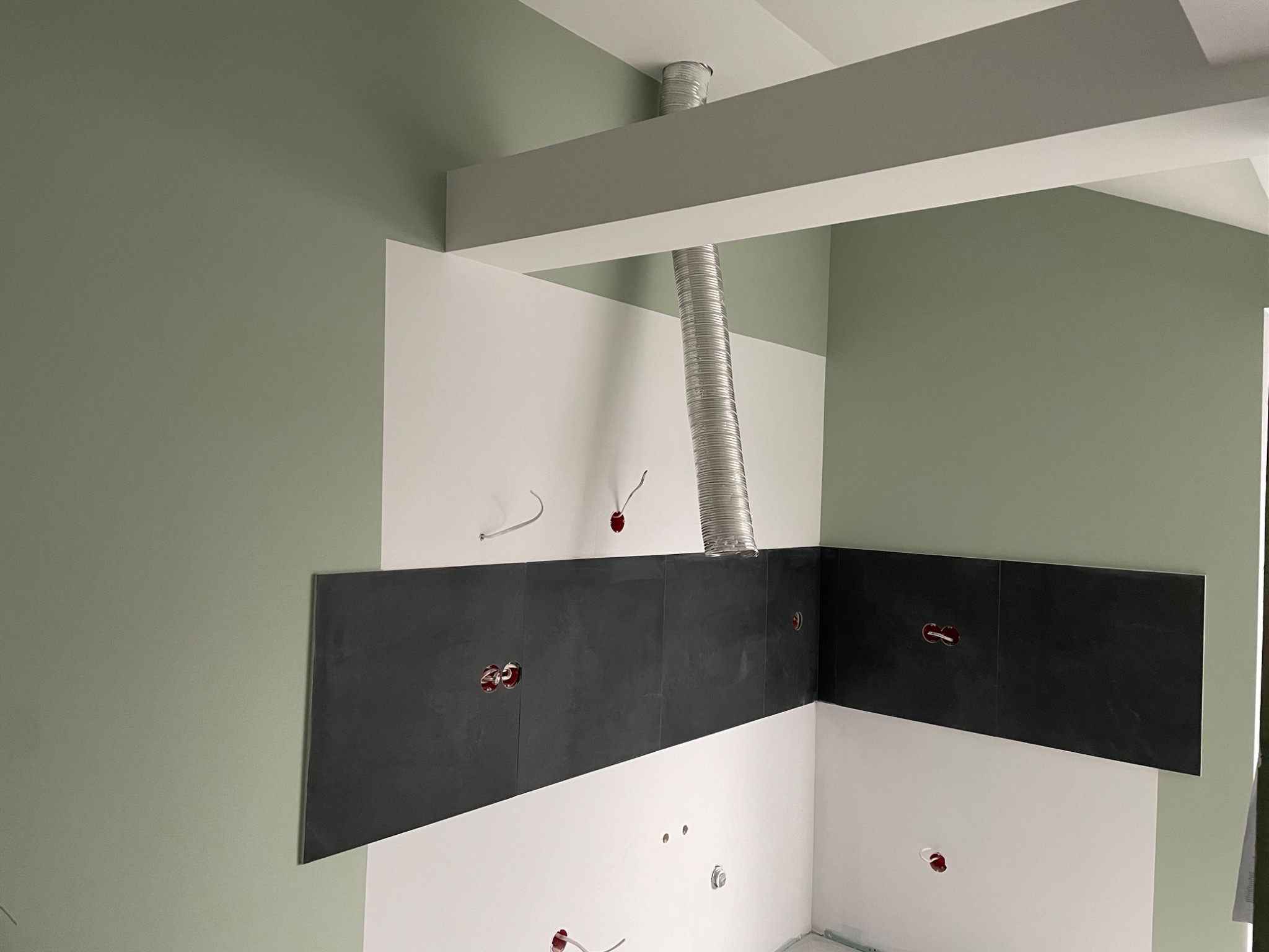 Wnętrze kuchni w trakcie prac wykończeniowych: położone ciemne płytki na ścianie, widoczne puszki elektryczne, ściany pomalowane na biało i oliwkowo, zamontowany kanał wentylacyjny.