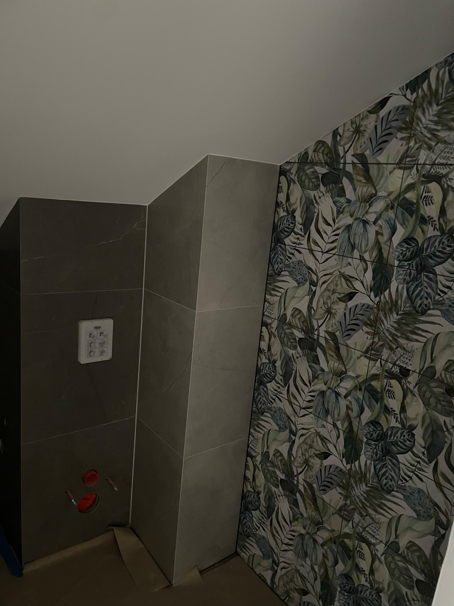 Fragment łazienki w trakcie remontu, z widocznymi płytkami w odcieniach szarości i zieleni z motywem roślinnym na jednej ze ścian, oraz szarą ścianą z zamontowanym panelem sterowania...